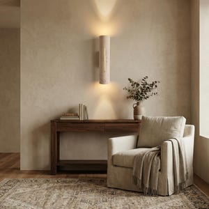 May include: A modern living room scene featuring a beige armchair with a throw blanket, a wooden console table with books, and a decorative wall sconce. The room has a neutral color palette with a patterned rug.