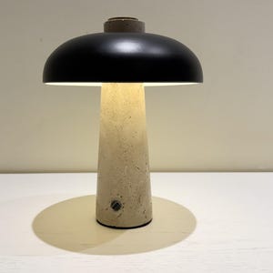 May include: A modern table lamp with a black lampshade and a beige concrete base. The lamp has a mushroom-like design, casting a warm glow. The base has a textured surface and a small switch.