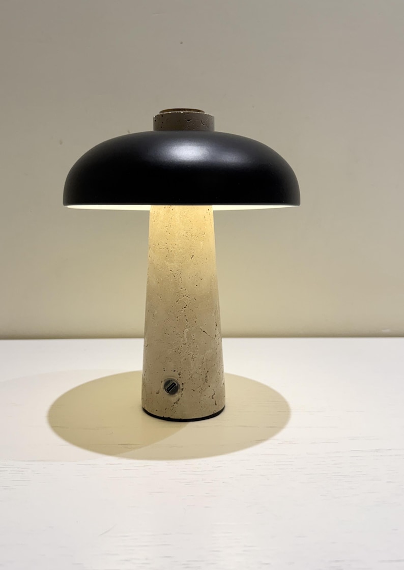 May include: A modern table lamp with a black lampshade and a beige concrete base. The lamp has a mushroom-like design, casting a warm glow. The base has a textured surface and a small switch.