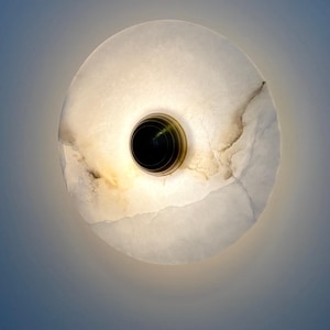 Può includere: Applique da parete rotonda bianca con un elemento centrale nero. L'applique è realizzata in un materiale traslucido, forse alabastro o marmo, con venature marroni. Il dispositivo ha un anello color ottone.