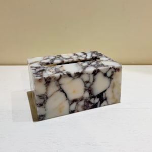 Può includere: Un portatovaglioli rettangolare in marmo bianco con venature grigio scuro e nere. La scatola ha un'apertura rettangolare sulla parte superiore per l'erogazione dei fazzoletti. È un oggetto decorativo per la casa.