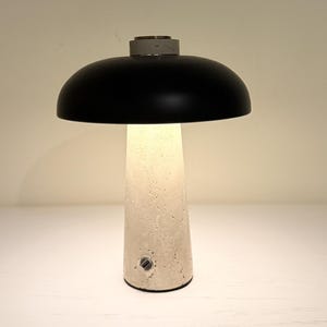May include: A modern table lamp with a black, dome-shaped shade and a textured, off-white base. The base has a conical shape and a small, circular power button. The lamp emits a warm, inviting glow.