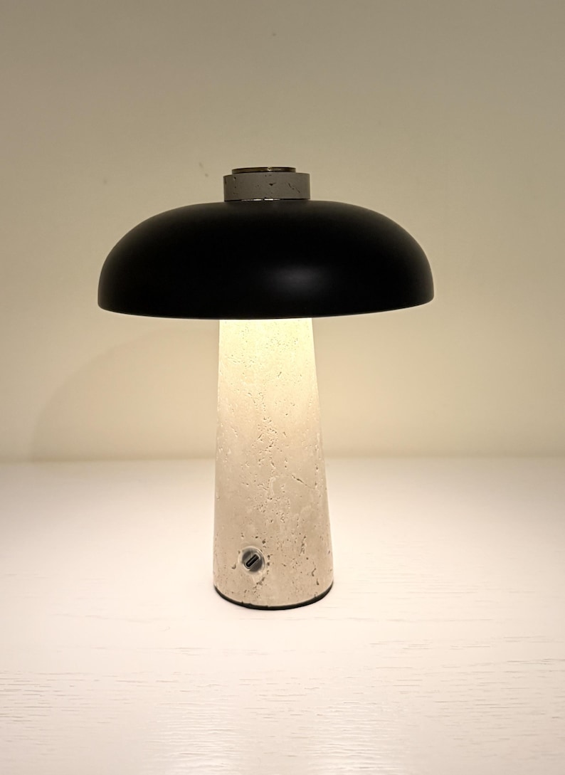 May include: A modern table lamp with a black, dome-shaped shade and a textured, off-white base. The base has a conical shape and a small, circular power button. The lamp emits a warm, inviting glow.