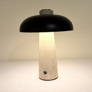 May include: A modern table lamp with a black dome-shaped shade and a textured, off-white base. The lamp has a cylindrical base with a small power port. The lamp is lit, emitting a warm glow.