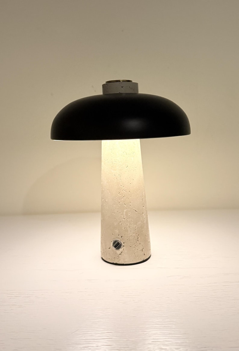 May include: A modern table lamp with a black dome-shaped shade and a textured, off-white base. The lamp has a cylindrical base with a small power port. The lamp is lit, emitting a warm glow.