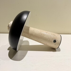 May include: A mushroom-shaped decorative object with a black top, white underside, and a beige, textured stem. The stem has a small, circular metal detail near the base. The object casts a shadow on a white surface.