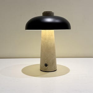 May include: A modern table lamp with a black lampshade and a beige, textured base. The lamp has a cylindrical base that widens towards the top, and a small, circular switch. The lamp is on a white surface.