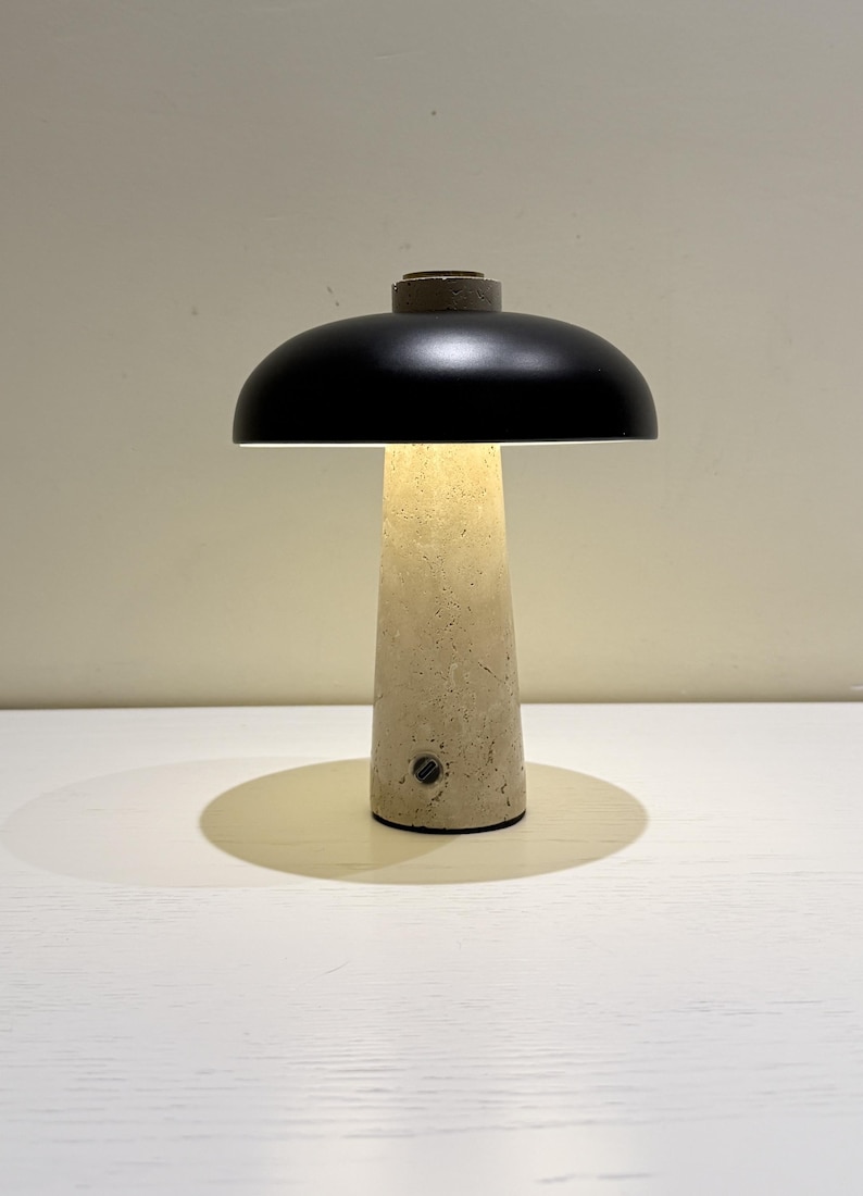 May include: A modern table lamp with a black lampshade and a beige, textured base. The lamp has a cylindrical base that widens towards the top, and a small, circular switch. The lamp is on a white surface.