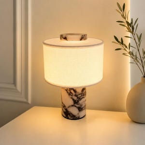 May include: A table lamp featuring a cylindrical base made of white and black marbled stone, topped with a circular white fabric drum shade and a matching marble accent piece at the very top, emitting a warm, soft glow from within the shade.