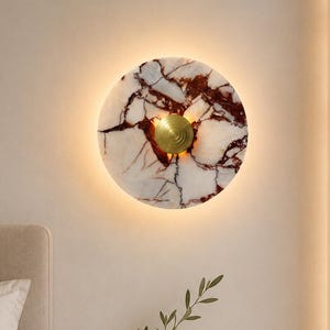 May include: A circular wall lamp featuring a white marble disc with prominent dark red veining. A small, gold-toned, ribbed circular fixture is centered on the marble, emitting a warm, soft glow from behind the disc onto the wall surface.