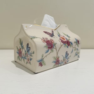 Pode incluir: Caixa de lenços de cerâmica creme com um desenho de borboletas e flores. A caixa tem formato de casa e possui uma borda dourada. O padrão floral inclui flores rosa, vermelhas e azuis com folhas verdes.