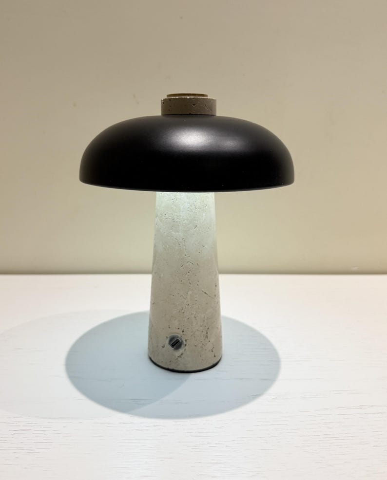 May include: A modern table lamp with a black dome-shaped shade and a concrete base. The lamp has a cylindrical base with a textured surface and a small switch. The lamp's design is minimalist and contemporary.