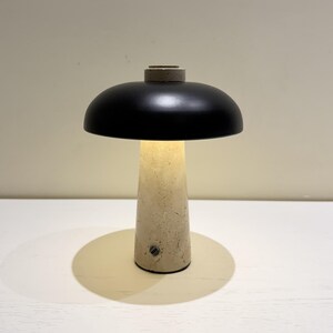 May include: A modern table lamp with a black, dome-shaped shade and a beige, conical base. The lamp is illuminated, casting a warm glow. The base appears to be made of concrete or a similar material. The lamp is on a white surface.