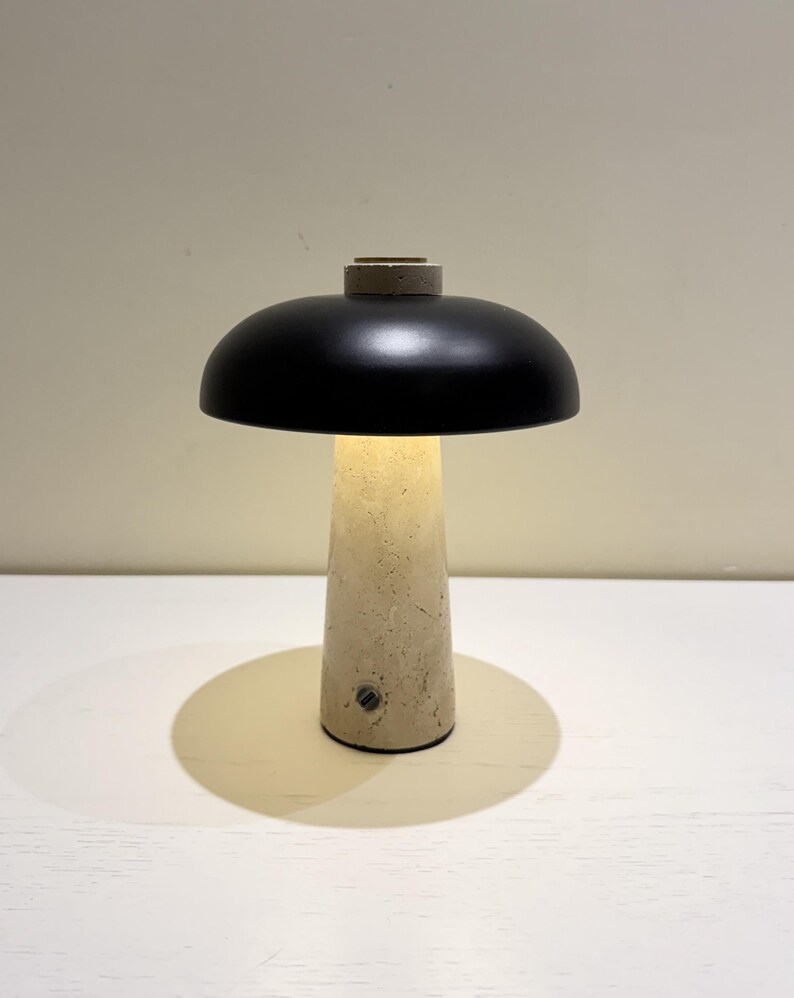 May include: A modern table lamp with a black, dome-shaped shade and a beige, conical base. The lamp is illuminated, casting a warm glow. The base appears to be made of concrete or a similar material. The lamp is on a white surface.