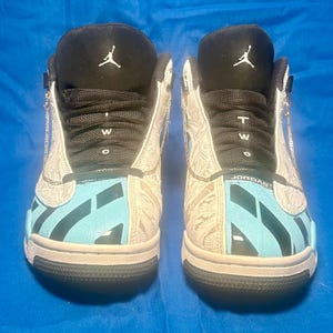 May include: Pair of high-top sneakers with a black and white design. The sneakers feature a light blue and black pattern on the toe and a white paisley design on the sides. The tongue is black with a white logo.