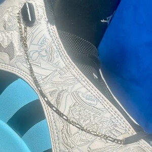 May include: Close-up of a white and light blue sneaker with intricate embossed designs. A silver chain dangles from the top of the shoe. The shoe has a black collar and a blue sole.
