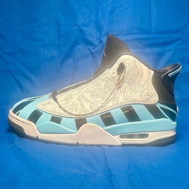 May include: A high-top sneaker with a light blue and black striped design on the lower portion. The upper part of the shoe is white with a detailed pattern. The shoe has a black tongue and heel, with a white sole.