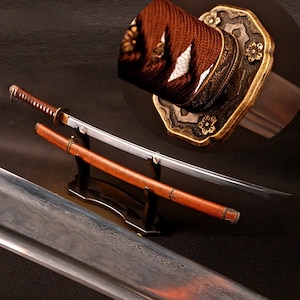 May include: A katana sword with a silver blade featuring a visible folded steel pattern, a brown cord-wrapped handle, and a brass-toned tsuba guard with floral engravings. The sword is displayed with a matching brown wooden scabbard on a black stand.