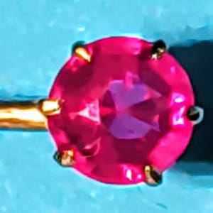 Op de afbeelding: Close-up van een goudkleurige oorbel met een gefacetteerde, ronde, felroze edelsteen. De edelsteen wordt op zijn plaats gehouden door vier kleine gouden pootjes. De oorbelpen is zichtbaar en strekt zich naar links uit.