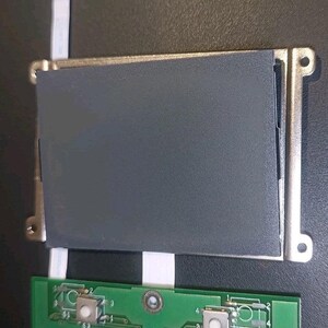 Può includere: Un modulo display elettronico rettangolare con schermo grigio scuro incorniciato in argento metallizzato, collegato tramite cavi a nastro bianchi a un circuito stampato verde con due pulsanti bianchi e il testo stampato 71-11A0S-D01.