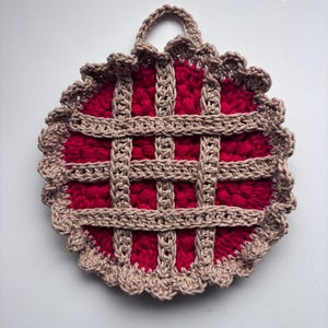 May include: A handmade crochet pie-shaped hot pad. The center is a vibrant red, with a lattice crust in a neutral tan color. The edge is scalloped, and a loop is attached for hanging. A charming kitchen accessory.