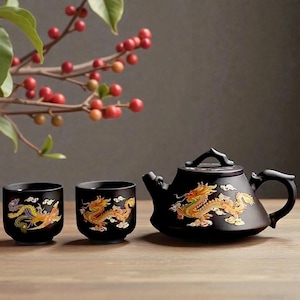Op de afbeelding: Zwarte keramische theeset met drakenontwerp. De set bevat een theepot en twee kopjes. De draak is afgebeeld in goud, geel en rood. De theeset staat op een houten oppervlak.