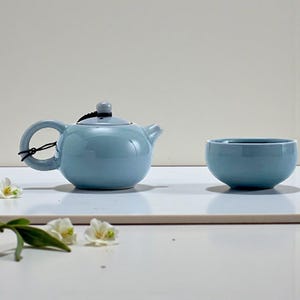 Może przedstawiać: Jasnoniebieski ceramiczny imbryk z pasującą pokrywką i małym uchwytem, obok pasującej filiżanki do herbaty. Imbryk ma mały dzióbek i czarny sznurek zawiązany wokół uchwytu. Białe kwiaty są rozrzucone wokół zestawu do herbaty.