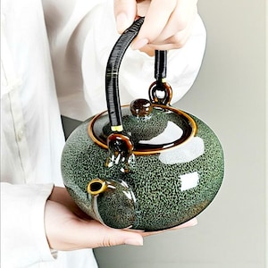 Könnte beinhalten: Eine grüne Keramik-Teekanne mit einem gesprenkelten Muster und einem schwarzen Griff. Die Teekanne hat einen runden Körper, einen Ausguss und einen Deckel mit einem kleinen Knopf. Der Griff besteht aus gewebtem Material. Die Teekanne wird von einer Person gehalten.