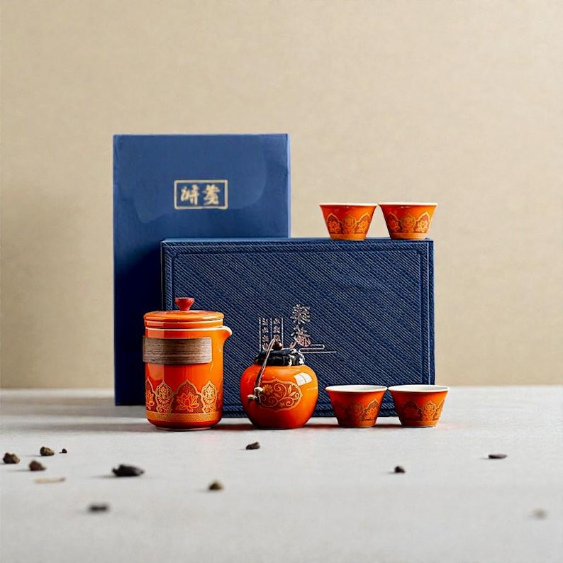 Peut inclure: Un service &agrave; th&eacute; en c&eacute;ramique orange avec des motifs floraux dor&eacute;s, comprenant une th&eacute;i&egrave;re, des tasses et un petit r&eacute;cipient d&eacute;coratif. L'ensemble est pr&eacute;sent&eacute; avec une bo&icirc;te bleue et un livre bleu avec du texte dor&eacute;.