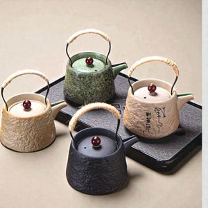 Peut inclure: Quatre théières en céramique de couleurs beige, verte et noire. Chacune a une anse tressée, un petit bec verseur et un couvercle avec un bouton rouge. Les théières sont disposées sur un plateau gris foncé.