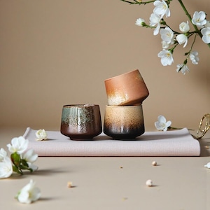 May include: Three ceramic cups with a gradient colour scheme, ranging from brown to black, are stacked on a pink cloth. The cups have a rustic, handmade appearance. White flowers and a gold ring are in the background.