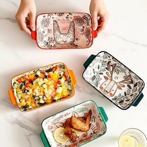 Puede incluir: Fuentes rectangulares de cerámica para hornear con diseños coloridos. Una fuente presenta un conejo, otra un ciervo y una tercera contiene una verdura horneada. Las fuentes tienen asas y están decoradas con motivos florales. Las fuentes son en tonos naranja, verde y rojo.