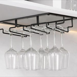 Puede incluir: Un estante de metal negro para copas de vino instalado debajo de un armario, que sostiene múltiples copas de vino transparentes. El estante está diseñado para sostener las copas boca abajo, mostrando su forma y claridad. Las copas están espaciadas uniformemente.