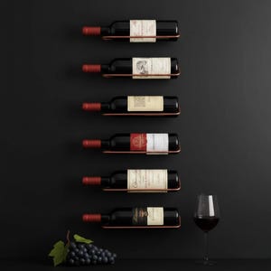 May include: Six wine bottles displayed on copper-coloured wall-mounted racks against a black background. A wine glass with red wine and a bunch of grapes with a green leaf are also present. The bottles have white labels.