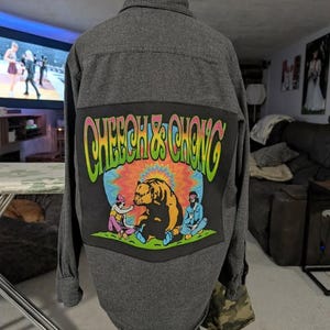 May include: A grey button-down shirt with a large patch on the back. The patch displays the text "Cheech & Chong" in colourful, psychedelic lettering, along with an illustration of a bear and two figures.
