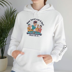 Puede incluir: Sudadera con capucha blanca con bolsillo delantero y capucha con cordón. La parte delantera presenta un gráfico colorido de una mujer jugando al billar con el texto "How hard can it be? Boys do it." La manga tiene el texto "Dawnzilla."