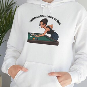 Puede incluir: Sudadera con capucha blanca con bolsillo delantero y cordón. El gráfico en la parte delantera presenta a una mujer jugando al billar con el texto "Confidence level: calling my shots."
