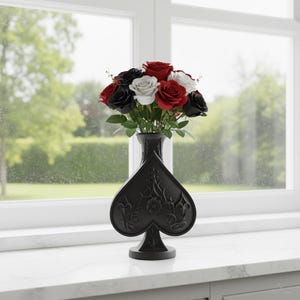 May include: A black vase in the shape of a spade, filled with red, white, and black roses. The vase has a detailed floral design and sits on a white windowsill in front of a window with a green outdoor view.