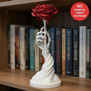 May include: A white sculpture of a skeleton hand holding a red rose. The base is round, and the stem is a twisted design. The rose is a deep red color. The sculpture is on a wooden shelf with books in the background.