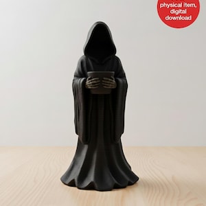 May include: A black Grim Reaper figurine holding a small, dark pot. The figure is draped in a hooded robe with long sleeves and a flared bottom. The figurine is on a light wood surface.