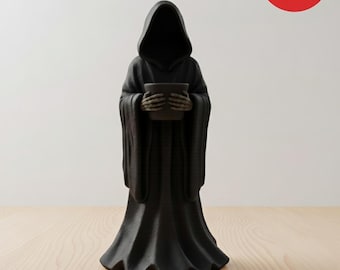 May include: A black Grim Reaper figurine holding a small, dark pot. The figure is draped in a hooded robe with long sleeves and a flared bottom. The figurine is on a light wood surface.
