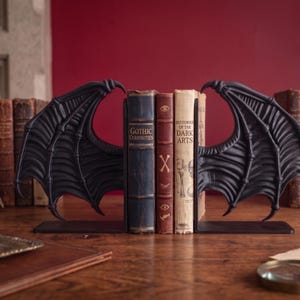 May include: Black metal bat wing bookends holding vintage books with titles like "Gothic Curiosities" and "Histories of the Dark Arts." The bookends are on a wooden surface, with a red background.