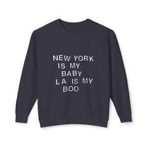 以下が含まれることがあります： 白い文字で「NEW YORK IS MY BABY LA IS MY BOO」と書かれたネイビーブルーのスウェットシャツ。長袖のクルーネックトップスはカジュアルなスタイルです。