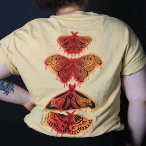 May include: Yellow t-shirt with the word "BUTTERMOTHS" in red at the top. The back of the shirt features four orange and black butterfly illustrations. The bottom of the shirt has the text "Label 001".