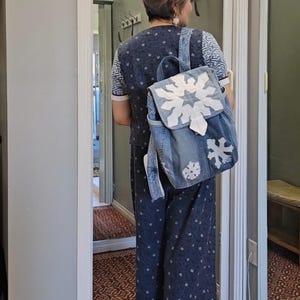 May include: A denim backpack with white snowflake appliqués is worn over a matching denim outfit. The backpack has a flap closure and adjustable straps. The outfit consists of a short-sleeved top and wide-leg trousers, both in a dark blue fabric with a small white pattern.