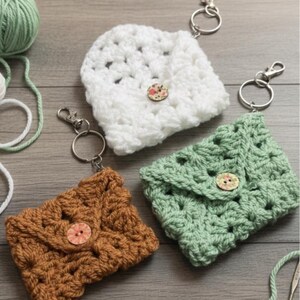 May include: Three crocheted keychains in white, brown, and green, each with a button closure and a silver-tone keyring. The handmade keychains are arranged on a wooden surface, with yarn and crochet hooks visible.