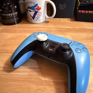 May include: A light blue and black video game controller with white accents. The controller is on a wooden surface. In the background are mugs, a small black device, and a Tetris arcade game.