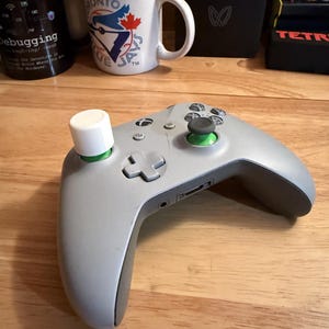 May include: A grey Xbox controller with white and green details. The controller features a white thumbstick extender and green joystick caps. It rests on a wooden surface.