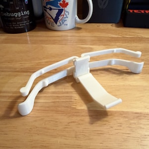 May include: A white 3D-printed device, possibly a phone or tablet holder, is centered on a wooden surface. The device has curved arms and a flat, angled support. A mug with the Toronto Blue Jays logo is in the background.