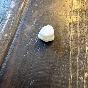 May include: A small, white, irregularly shaped object, possibly a decorative element or component, rests on a dark, textured wooden surface. The object has a layered appearance, with a slightly uneven top.